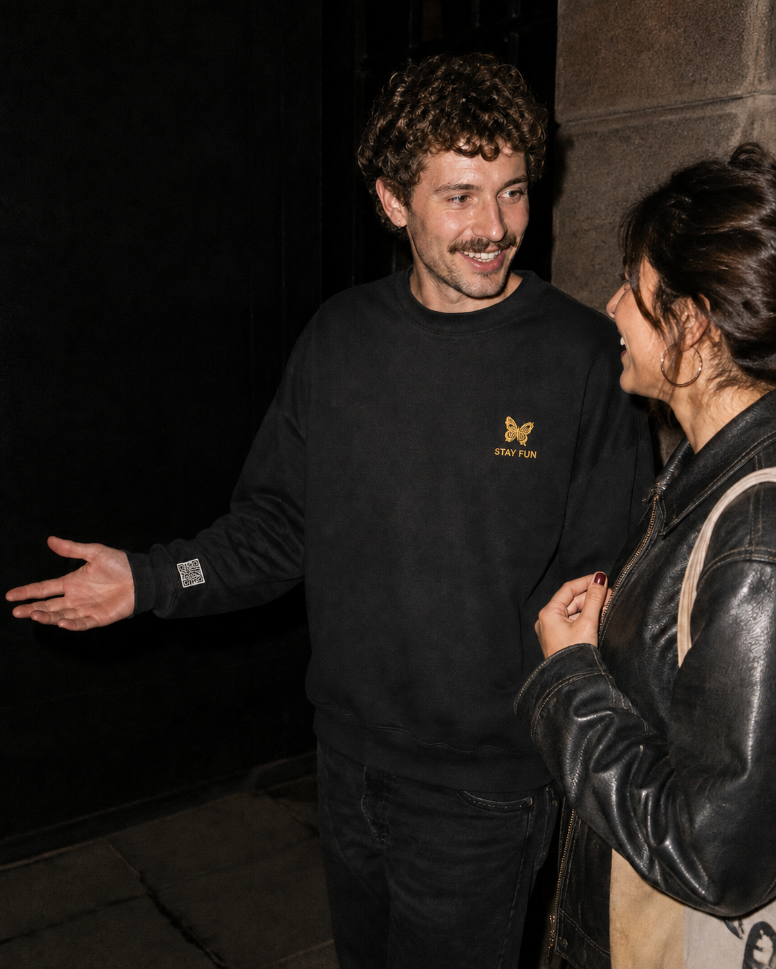 Two people talking outside at night; one wears a black stayfun sweatshirt with a small yellow butterfly and sleeve code.